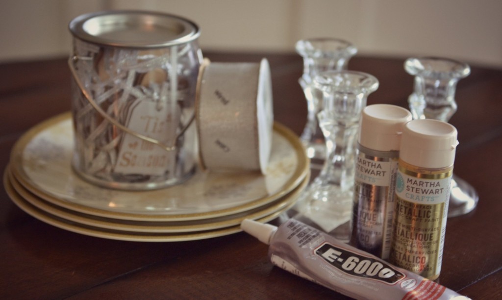 Assemble your materials for the DIY cake stand: holiday plates, glass candlesticks, Martha Stewart metallic paint, paint brush, E-6000 adhesive, clear cellophane, ribbon and gift tags. 