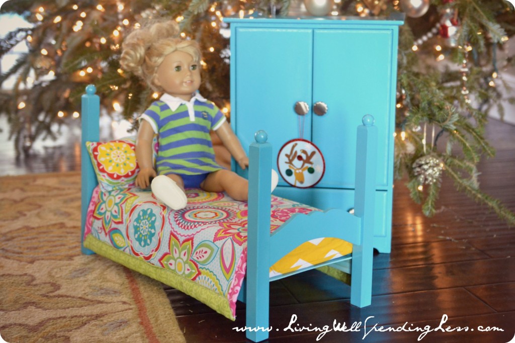 An American Girl doll sitting on a doll bed next to an armoire. 