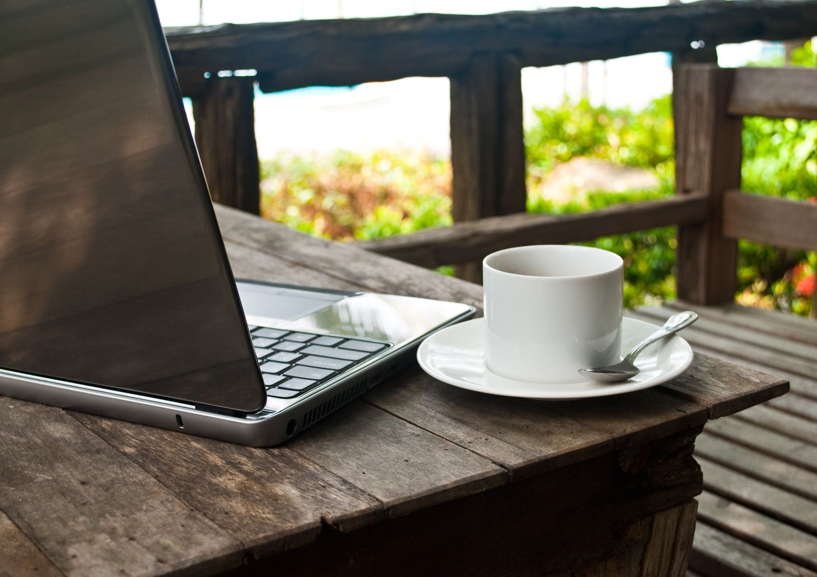 A cup on coffee and a laptop on top of a table outside on a wooden balcony. 