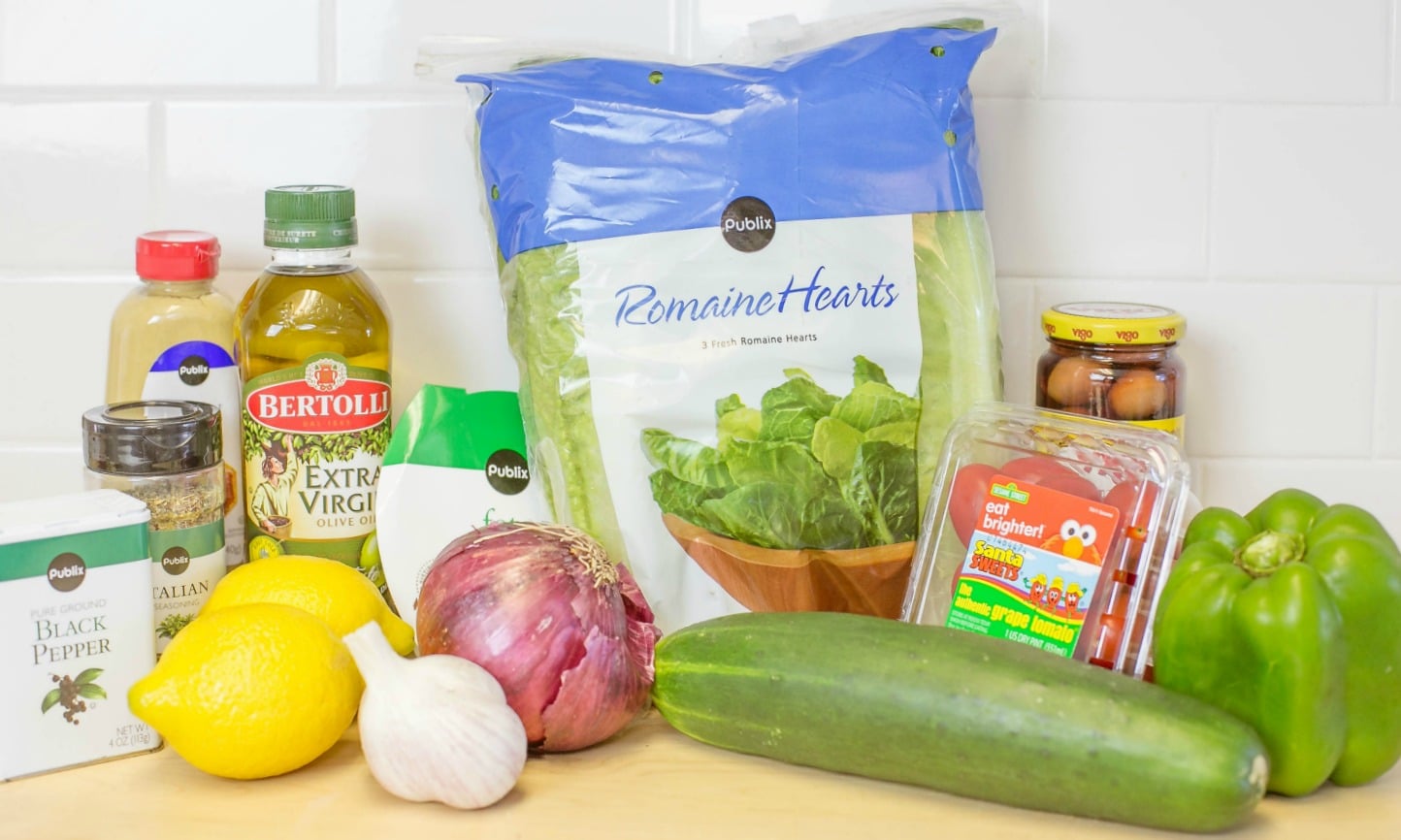 Assemble your Greek salad ingredients: Romaine hearts, black pepper, Italian seasoning, mustard, olive oil, lemons, garlic, red onion, cucumber, green pepper, tomatoes, olives and feta cheese. 