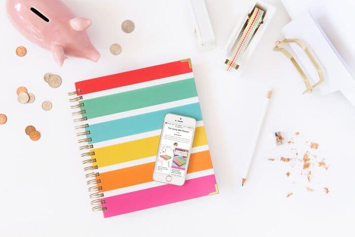 A colorful notebook, piggy bank, tape, pencil and smart phone on top of a white desk. 