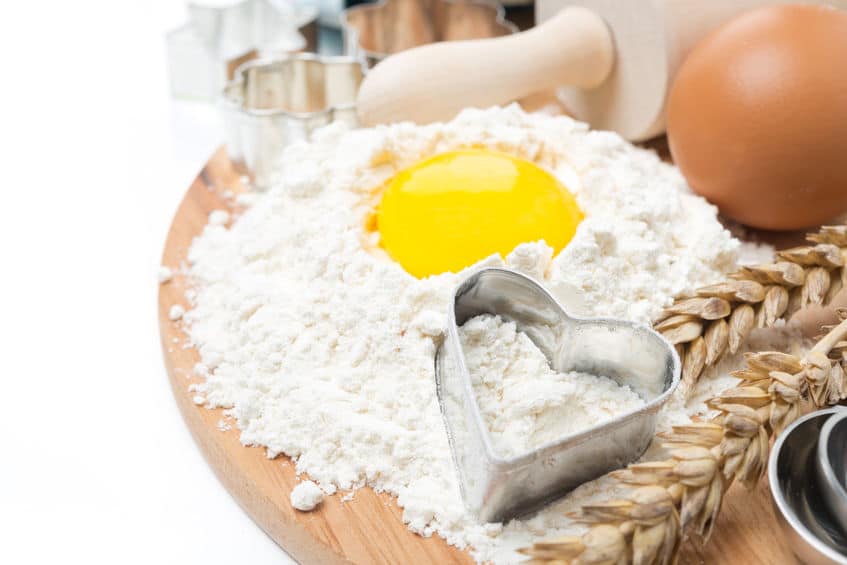 A wooden table with flower, an egg yolk, cookie cutters and a rolling pin. 