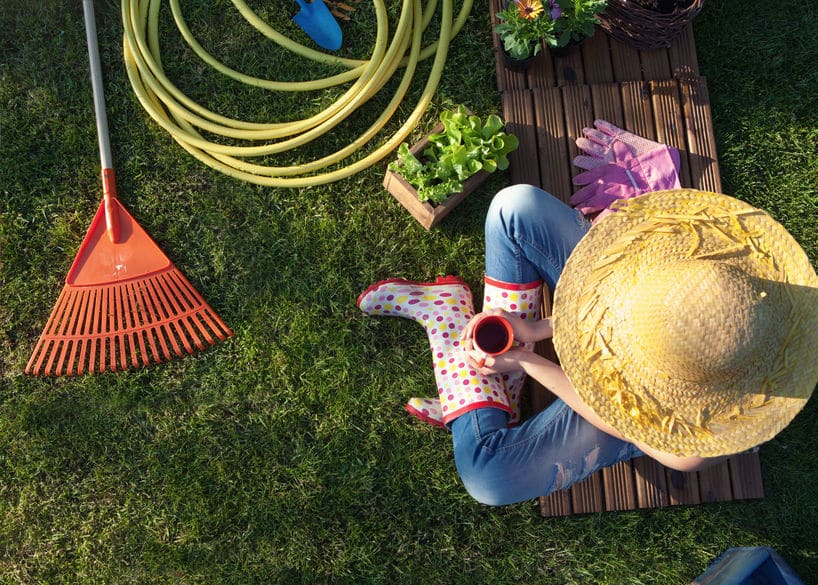 Woman wearing a sun hate and rain boots while sitting on a bench near gardening supplies like a rake, gloves and a hose. 