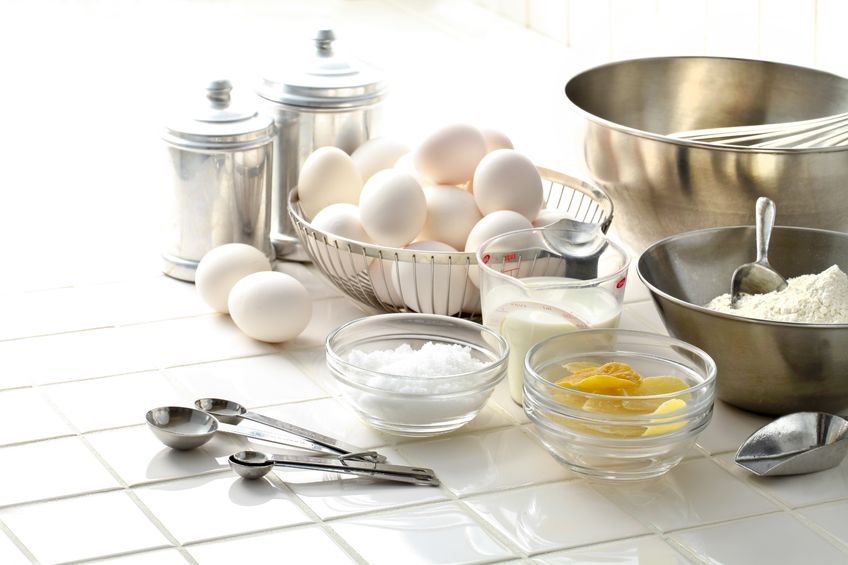 A kitchen counter with various utensils, bowls, a basket of eggs, a measuring cup with milk and other ingredients. 